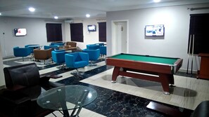 A billiards room with a pool table, blue and brown chairs, and two televisions mounted on the walls.