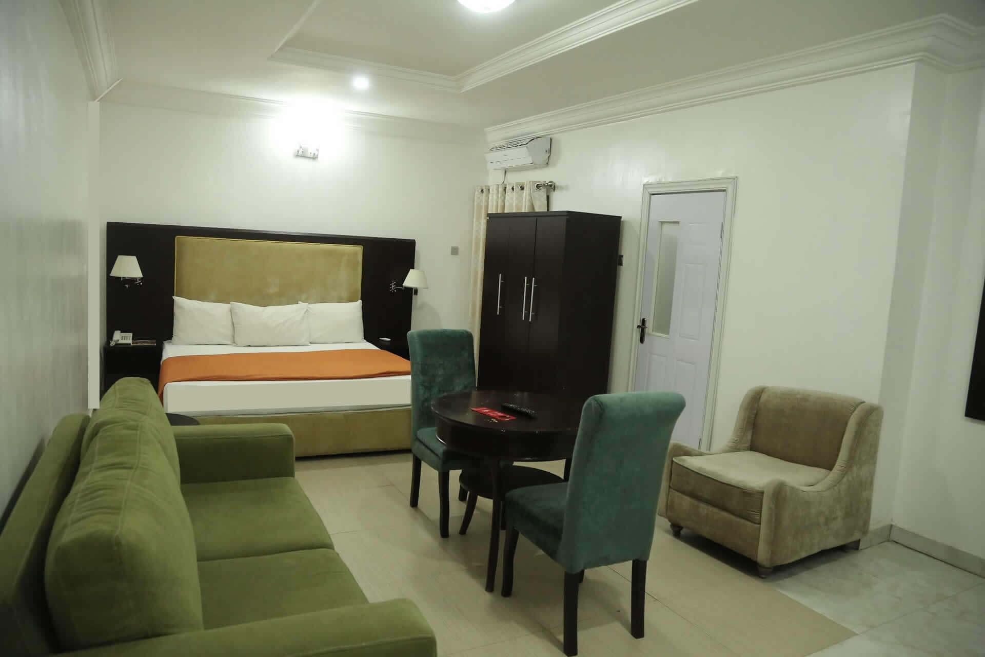 Habitación de hotel con una cama, un sofá verde, un sillón beige, una mesita y un armario.