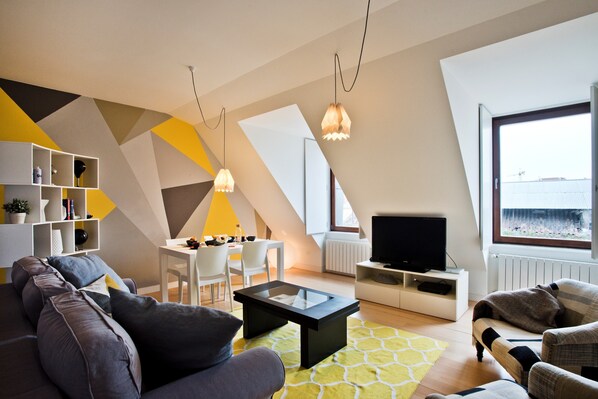 A modern living room with a sofa, coffee table, and TV, a dining area with chairs, and a large window with a view.