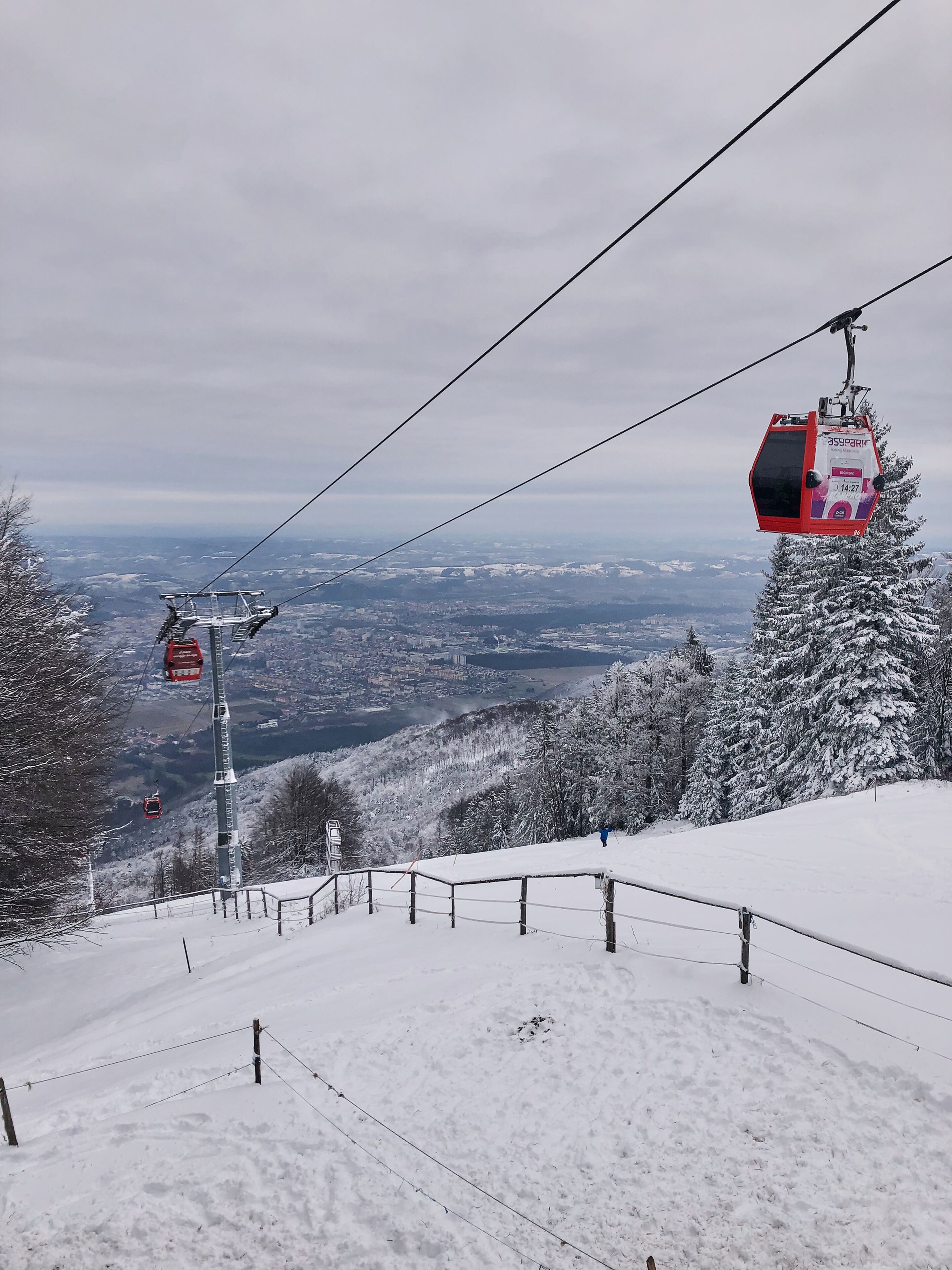 Un télésiège avec des cabines qui se déplacent le long du câble, un paysage enneigé et une vue lointaine sur une ville.