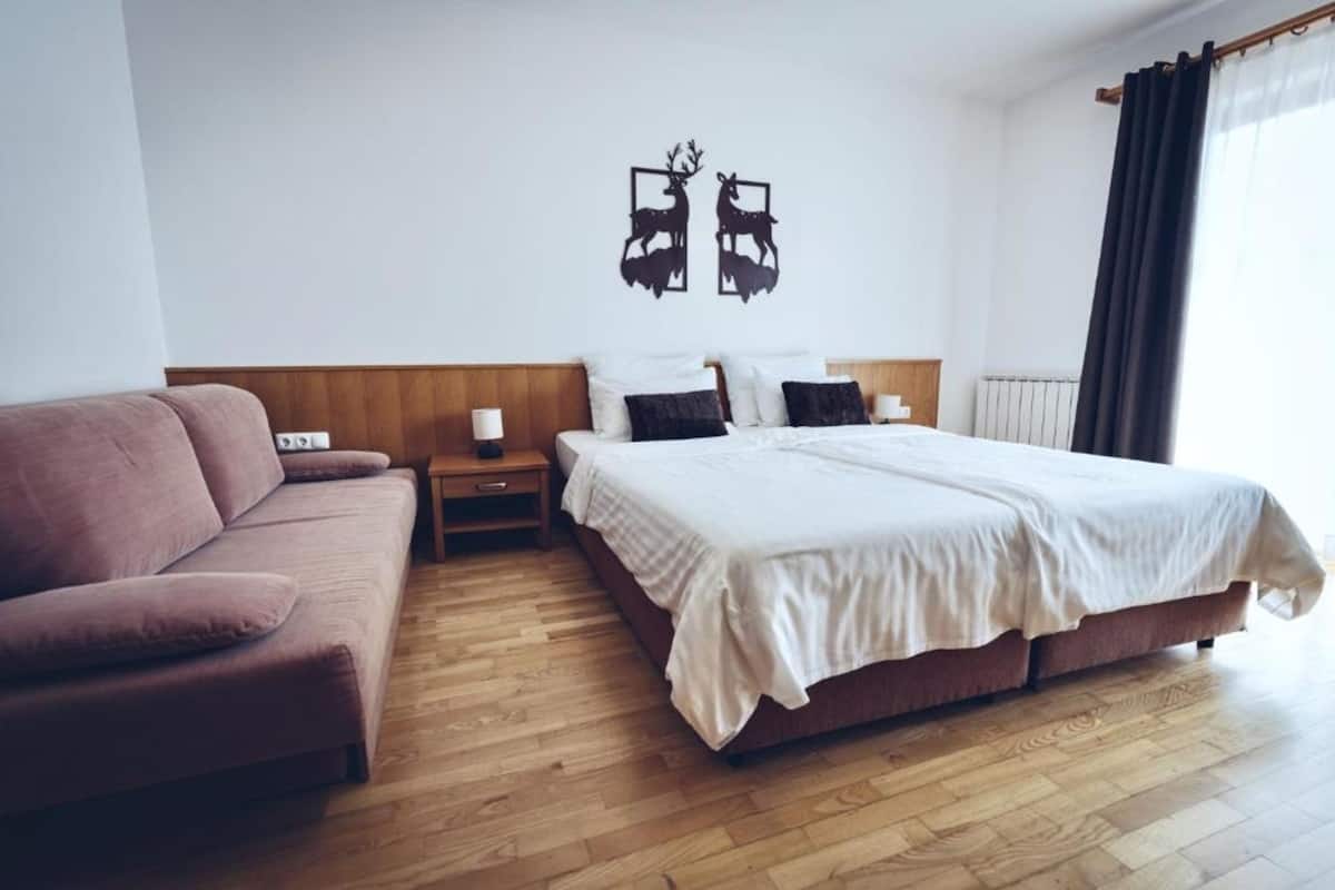 Ett hotellrum med trägolv, en säng med vita sängkläder, en brun soffa, ett nattduksbord och en väggdekoration med motiv av hjortar.