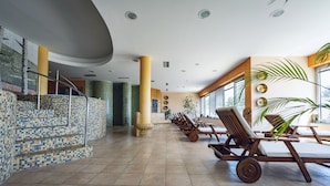 A spacious indoor area with tiled flooring, a staircase with mosaic tiles, wooden lounge chairs, and large windows allowing natural light. - Pohorje Village Wellbeing Resort – Wellness & Spa Hotel Bolfenk (Maribor)