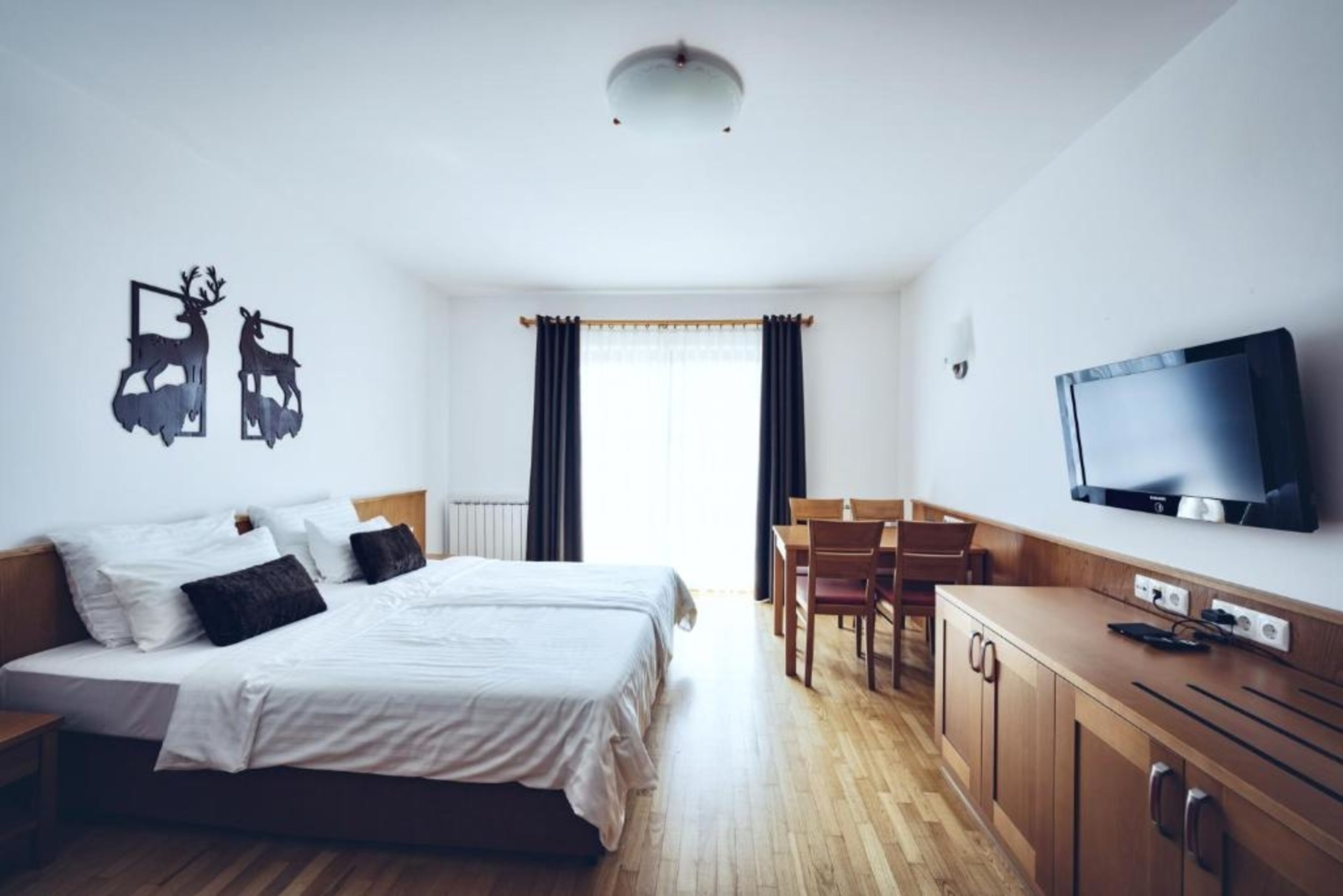 A hotel room with a bed, a TV, a dining table, and a window with curtains.