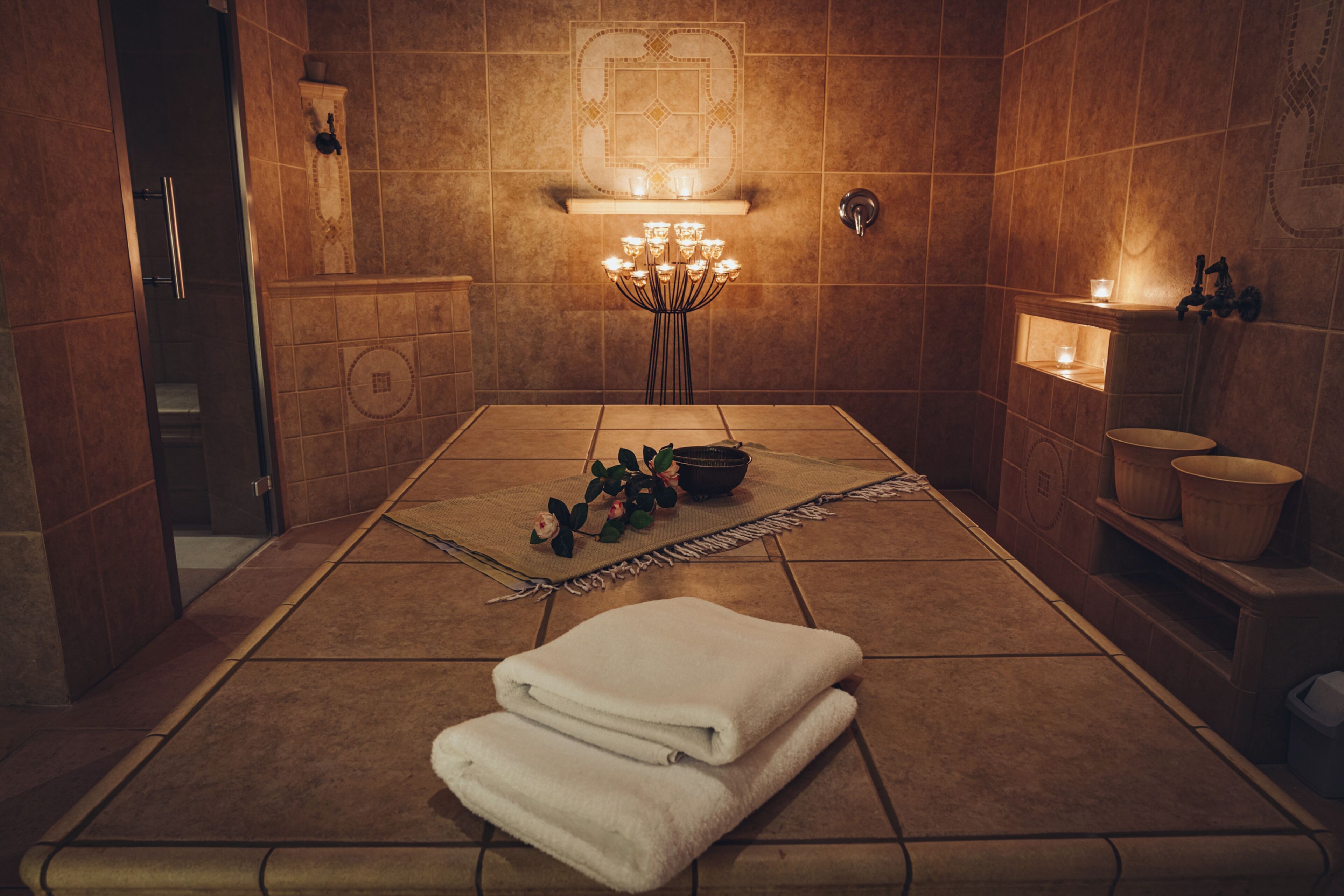 Un cuarto de spa con piso de baldosas, una camilla de masaje, una planta pequeña y un estante con velas.