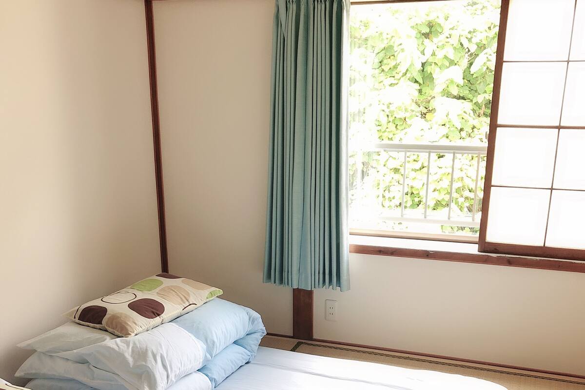 A room with two beds, a window with a curtain, and a view of greenery outside.