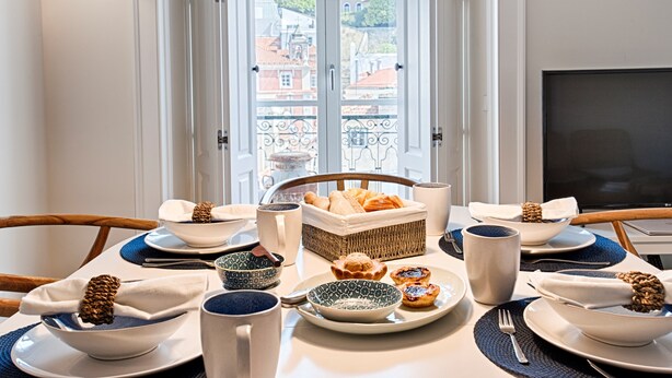 A dining table set with plates, bowls, and a basket of pastries, with a view of buildings through a window.