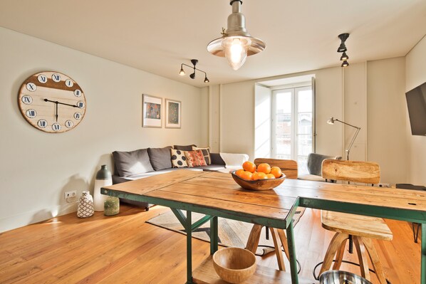 A modern living room with a wooden dining table, a bowl of oranges, and a wall clock.