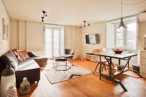 A modern living room with a sofa, dining table, and chairs, a television mounted on the wall, and a large window providing natural light.