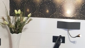 A white vase with green plants on a wooden table, a black and gold patterned wall, and a black electrical outlet on a white wall. - Waldhotel und Restaurants Lemberg (Hagen)