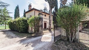 Une villa de style rustique, avec une allée de gravier menant à une entrée fermée par un portail, entourée d’arbres majestueux et sous un ciel dégagé.