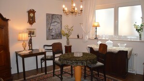 A dining area with a round table, chairs, a chandelier, a clock, and a window with curtains. - Ulrike's Pension am Kurpark (Bad Sooden-Allendorf)