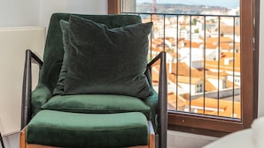 A green chair with a cushion, a black floor lamp, and a view of a cityscape through a large window with a metal railing.