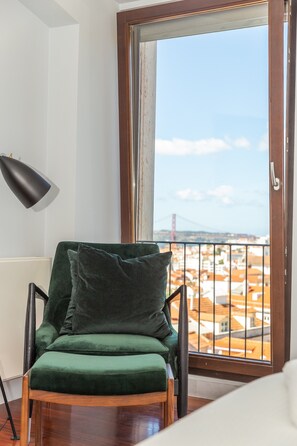 Une chaise verte avec un coussin, un lampadaire noir et une vue sur le paysage urbain, visible à travers une grande fenêtre dotée d’une rambarde en métal.