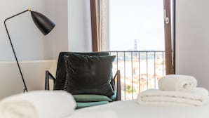 A hotel room with a bed, a chair with a green cushion, a black floor lamp, and a view of a bridge through the window.