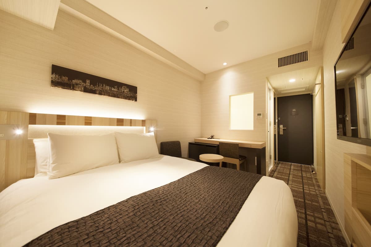 A hotel room with a large bed, a desk, a chair, a small table, and a mirror.