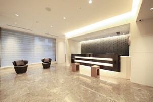 Una moderna recepción con suelo de mármol, dos sillones oscuros, un mostrador de recepción y una pared con patrón geométrico.