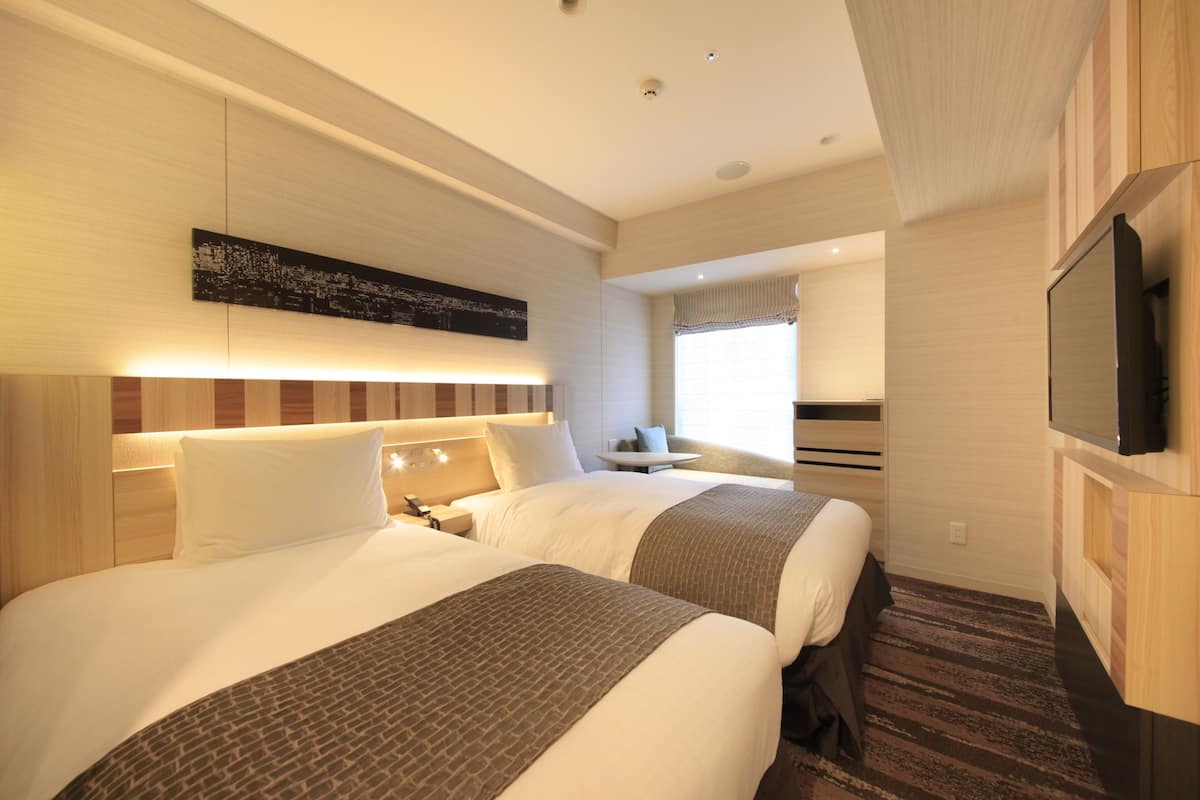 A hotel room with two beds, a flat-screen TV, a small table, and a window with curtains.