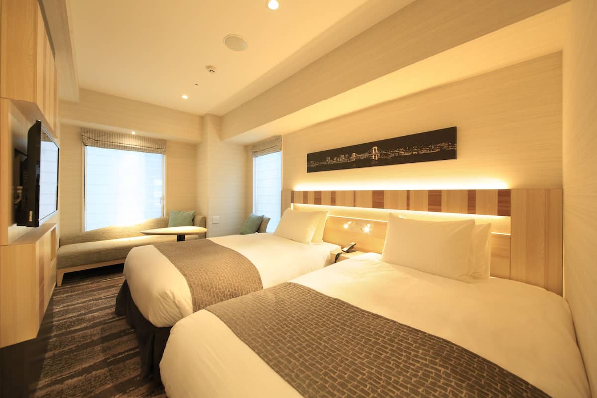 A hotel room with two beds, a flat-screen TV, a cityscape artwork, and a window with blinds.