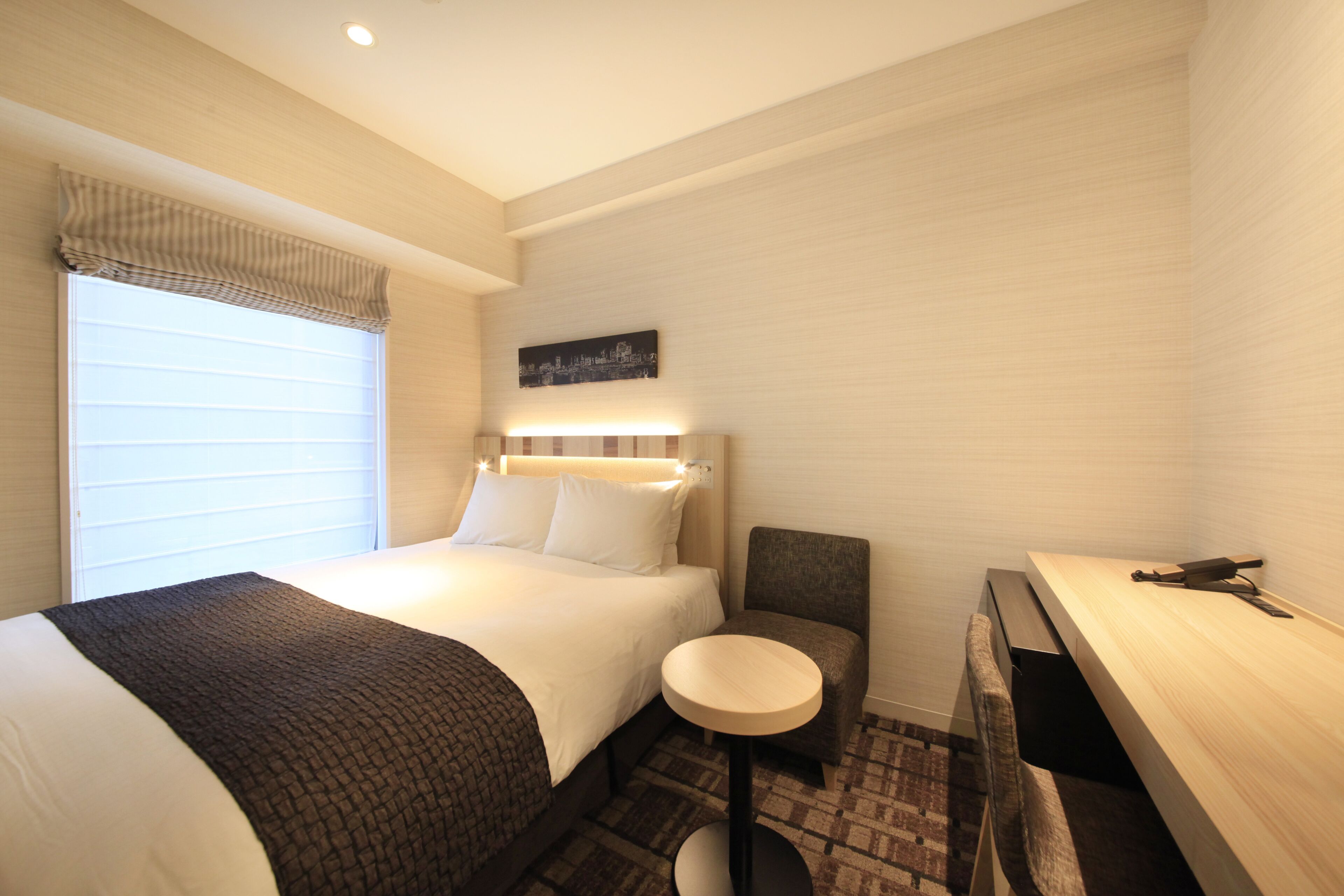 A hotel room with a large bed, a small round table, a chair, a desk, and a window with blinds.