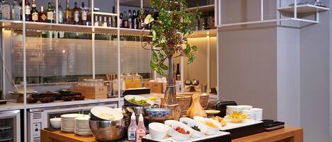 Montaje de buffet con diversos alimentos y condimentos sobre una mesa de madera, rodeado de estantes con botellas y copas al fondo.