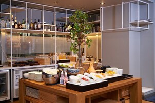 Montaje de buffet con diversos alimentos y condimentos sobre una mesa de madera, rodeado de estantes con botellas y copas al fondo.