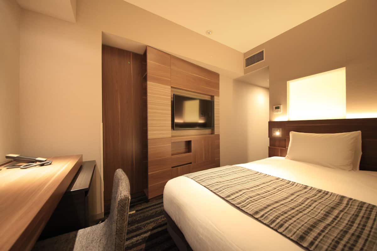 A hotel room with a bed, a desk, a chair, a television, and a wall-mounted light.