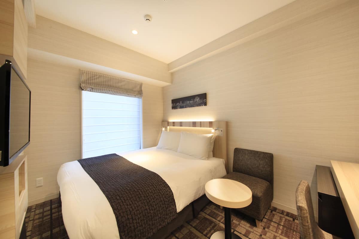 A hotel room with a large bed, a small round table, a chair, a television, and a window with blinds.