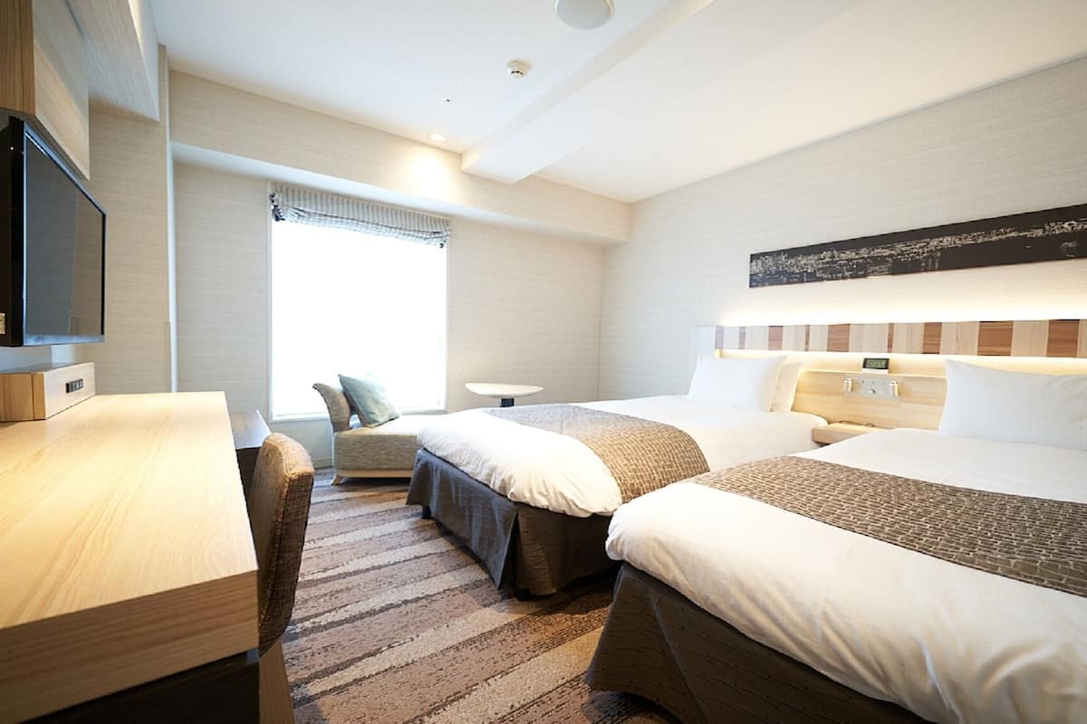 A hotel room with two beds, a desk, a chair, a TV, and a window with curtains.