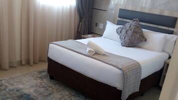 A hotel room with a large bed, a nightstand, a lamp, a window with curtains, and a patterned rug.