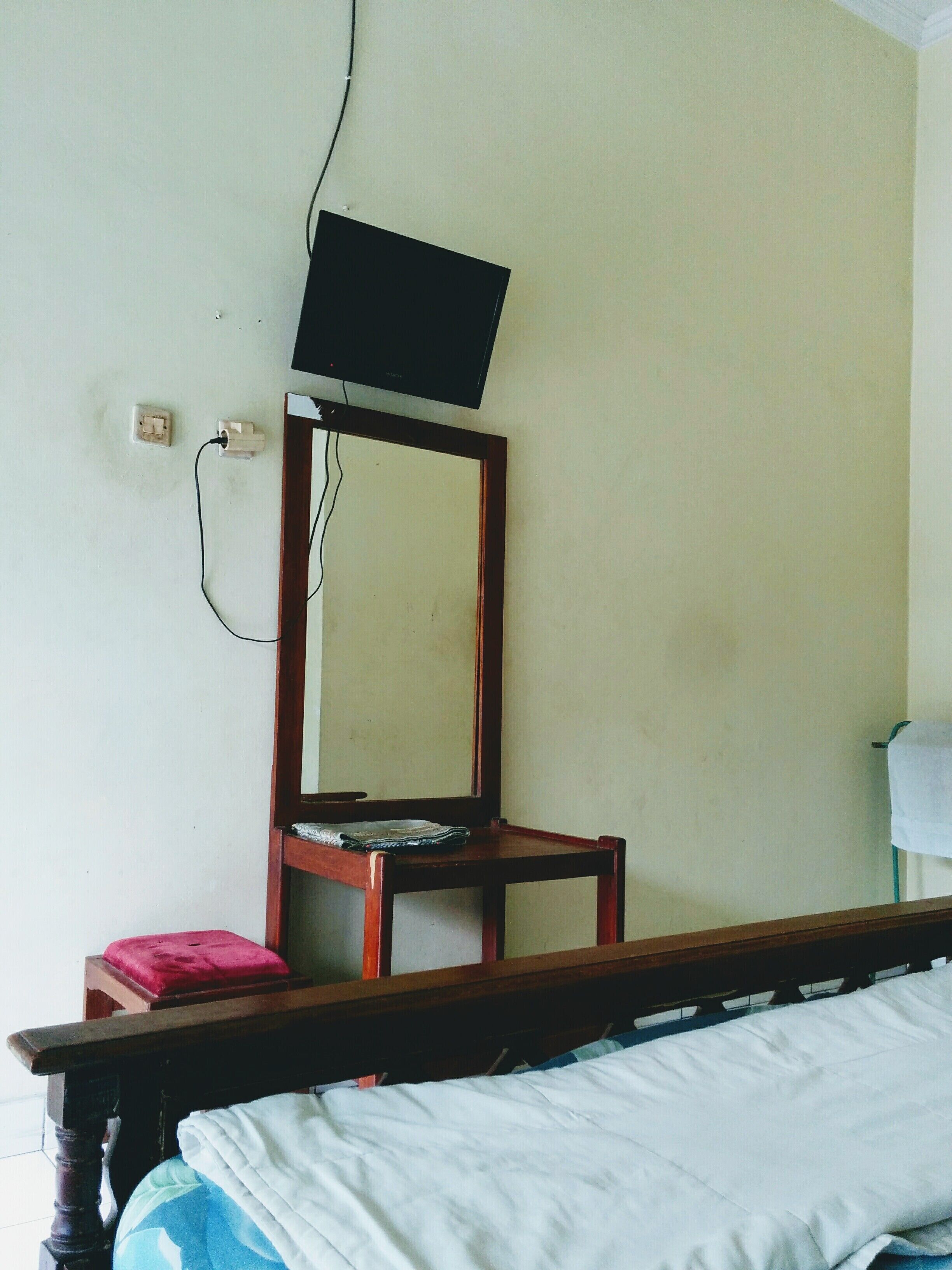 A bedroom with a wooden bed frame, a single wooden chair, a television mounted on the wall, and a small red cushion on the bed.