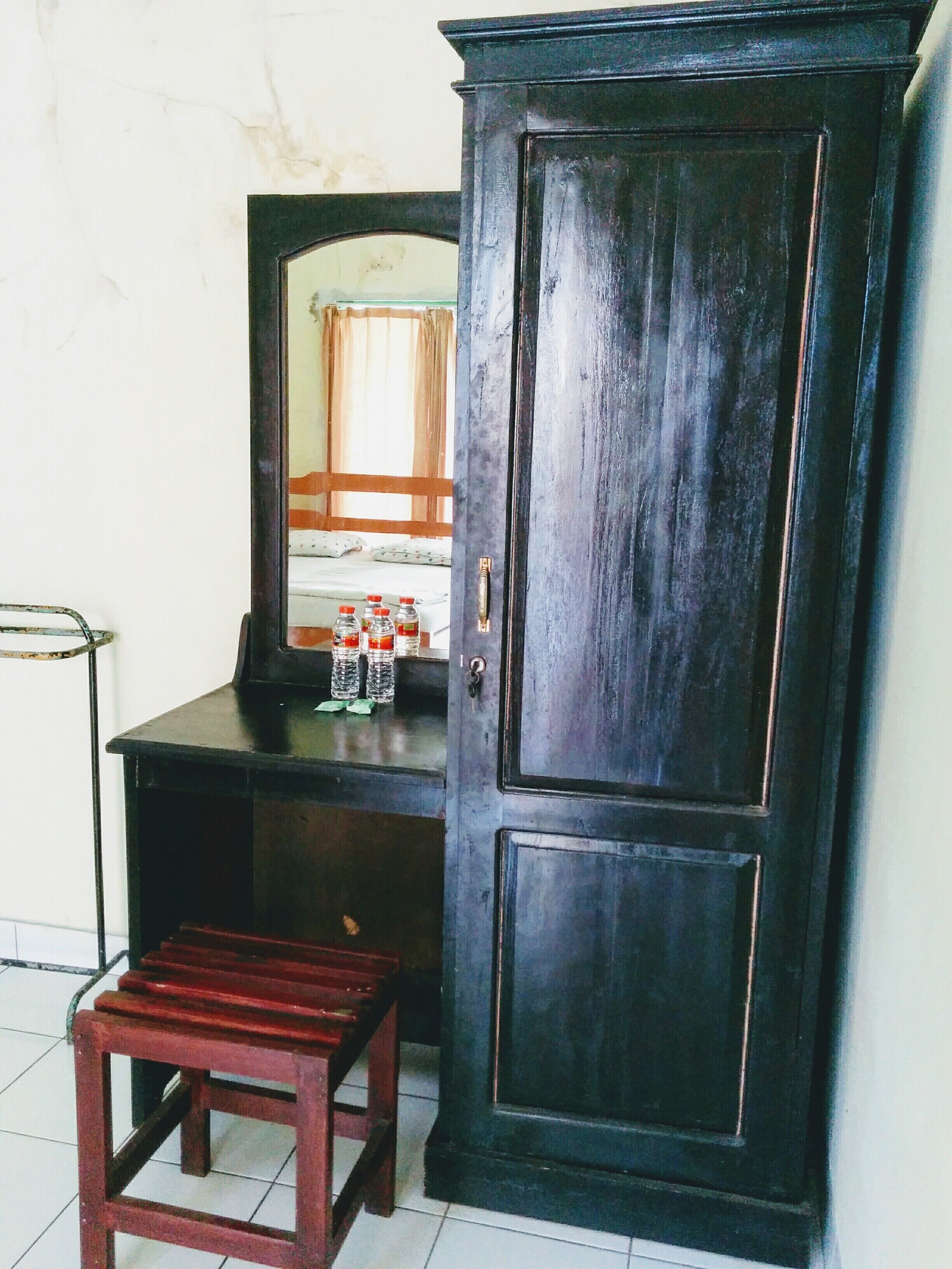A dark wooden wardrobe with a mirror, a small wooden stool, and a shelf with bottled water.
