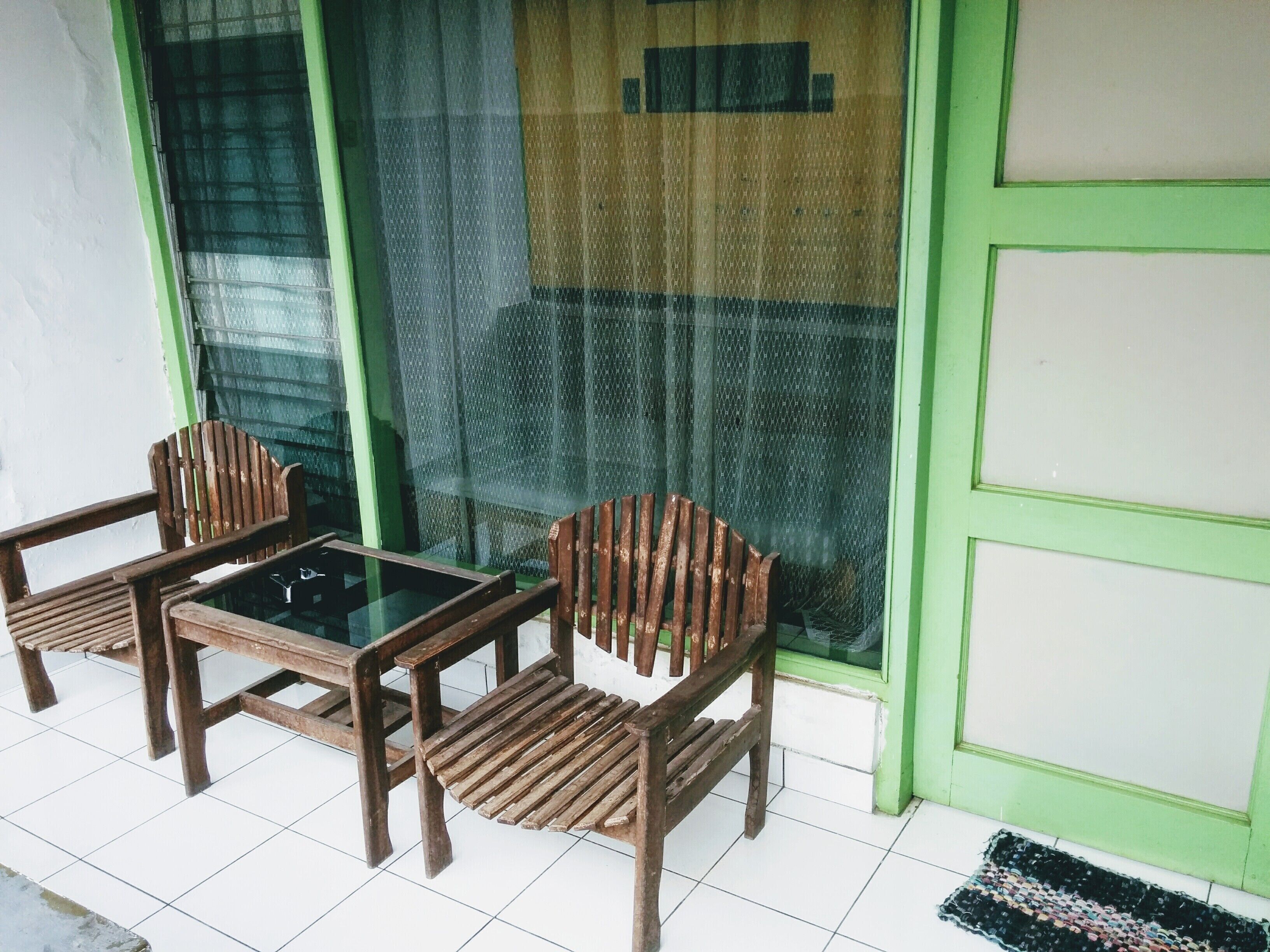 Un petit espace extérieur aménagé avec deux chaises en bois et une table en verre, une porte verte et une fenêtre munie d’un moustiquaire.