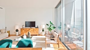 Un salon moderne doté d’une grande fenêtre, d’un téléviseur à écran plat, d’un meuble en bois et d’un coin salon avec un canapé et des fauteuils.