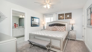 A bedroom with a bed, a bench, a nightstand, a ceiling fan, a mirror, and artwork on the walls. - Stunning Lakeview Townhome w/ Private Pool! (Kissimmee)