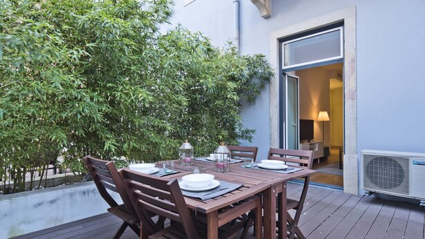 Una terraza de madera al aire libre con una mesa lista para comer, rodeada de sillas, y un árbol grande al fondo.