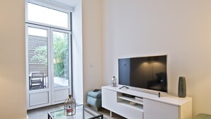 Una sala de estar moderna con un televisor de pantalla plana grande sobre un mueble blanco, una mesa de centro de vidrio y un amplio ventanal corredera que da a un área exterior con un banco y follaje.