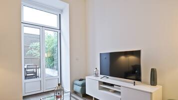 Uma sala de estar moderna com uma grande televisão de tela plana sobre um móvel branco, uma mesa de centro de vidro e uma porta de correr de vidro que dá para uma área externa com um banco e vegetação.
