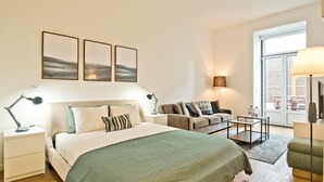 Un dormitorio moderno con una cama grande, un sofá, un escritorio y una lámpara. Hay tres cuadros enmarcados en la pared y una ventana con persianas.