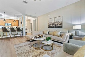 A modern living room with a sofa, coffee table, and chairs, a kitchen in the background, and artwork on the wall. - Cozy Vista Cay Condo! Free Resort Access (Orlando)