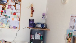 A room with a white floral sofa, a white chair, a wooden desk, a corkboard with various papers, a bookshelf, and a clock on the wall. - Guest House Ringo (Otaru)