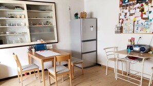 A room with a wooden dining table, two chairs, a refrigerator, a bulletin board with notes, and a window with shelves. - Guest House Ringo (Otaru)
