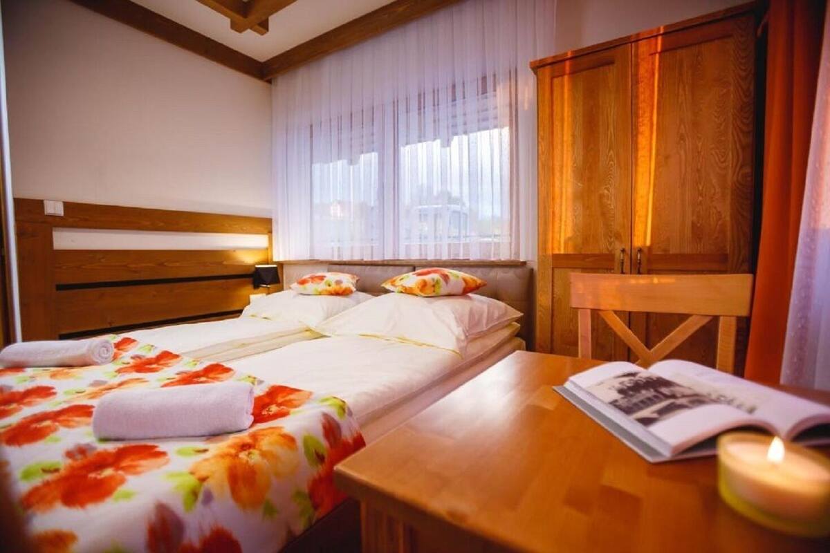 A hotel room with a wooden bed, white linens, a wooden nightstand, a wooden cabinet, and a window with sheer curtains.
