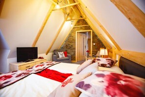 Ein gemütliches Schlafzimmer im Dachgeschoss mit schrägen Wänden, einem Bett mit floralem Bettzeug, einem Fernseher, einem kleinen Tisch mit Lampe und einem Sessel.