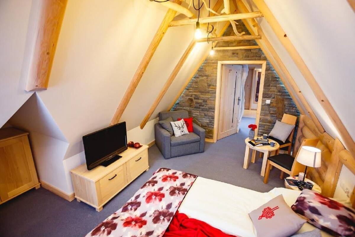 A cozy attic bedroom with a bed, a TV, a sofa, and a small table with a lamp.