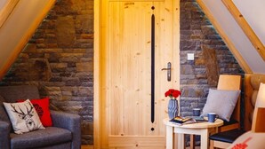 Une pièce chaleureuse avec un mur en pierre, une porte en bois, une petite table sur laquelle se trouvent un livre et un vase rempli de fleurs rouges, et une chaise agrémentée d’un coussin rouge.