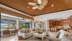 Uma sala de estar espaçosa com um sofá seccional grande, uma mesa de centro e uma área de jantar com banquetas. O ambiente possui teto de madeira e uma grande janela que oferece vista para o exterior.