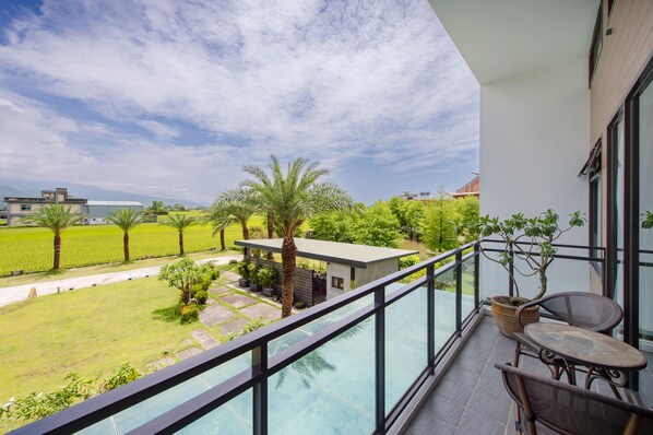 A balcony with a glass railing, a table, and chairs, overlooking a garden with palm trees and a modern building in the background.