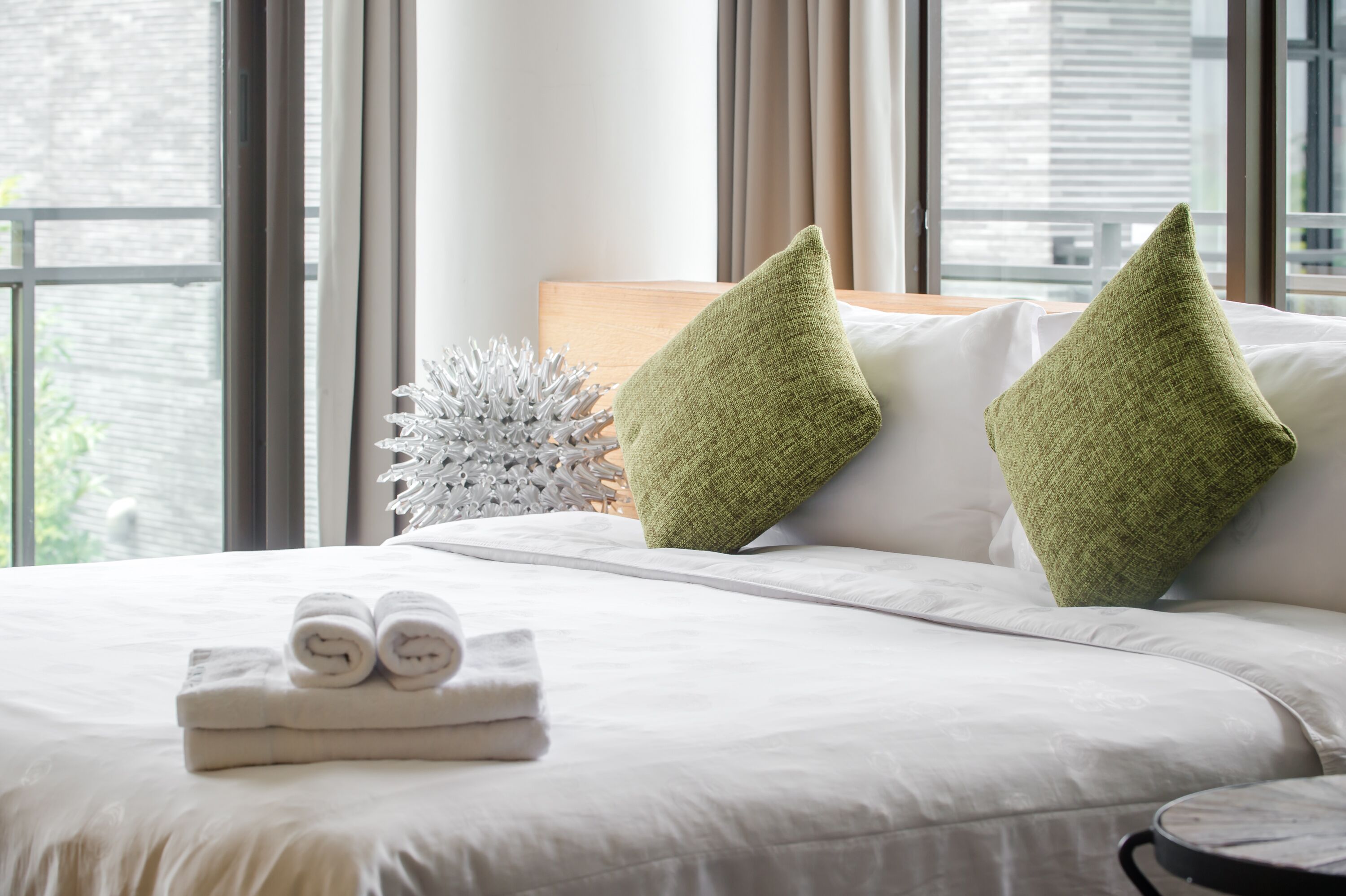 Cama bem arrumada com roupa de cama branca e almofadas verdes, uma planta branca decorativa e uma pilha de toalhas dobradas.