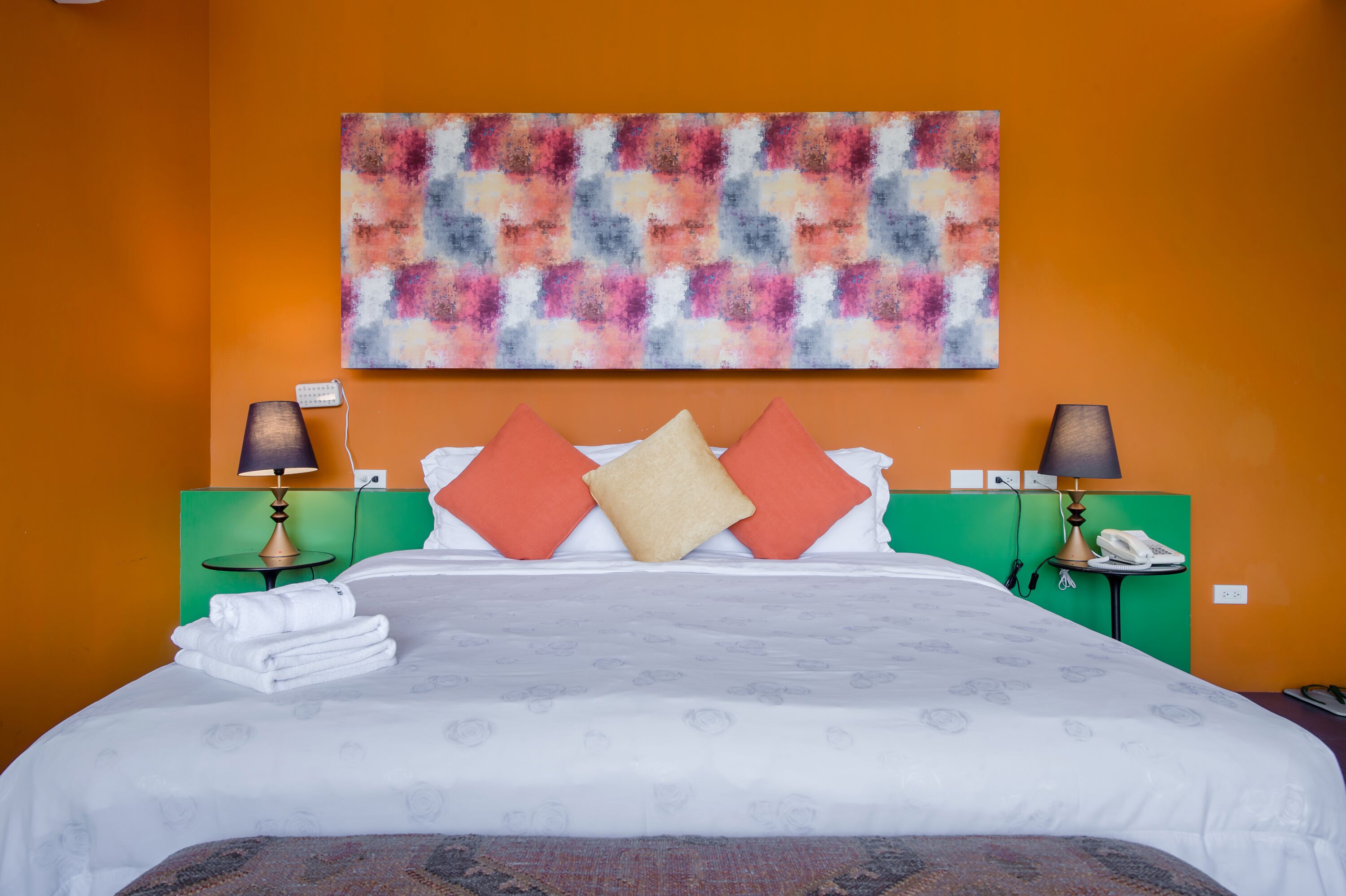 A hotel room with a bed, two nightstands, a lamp, a phone, and a colorful abstract painting on the wall.