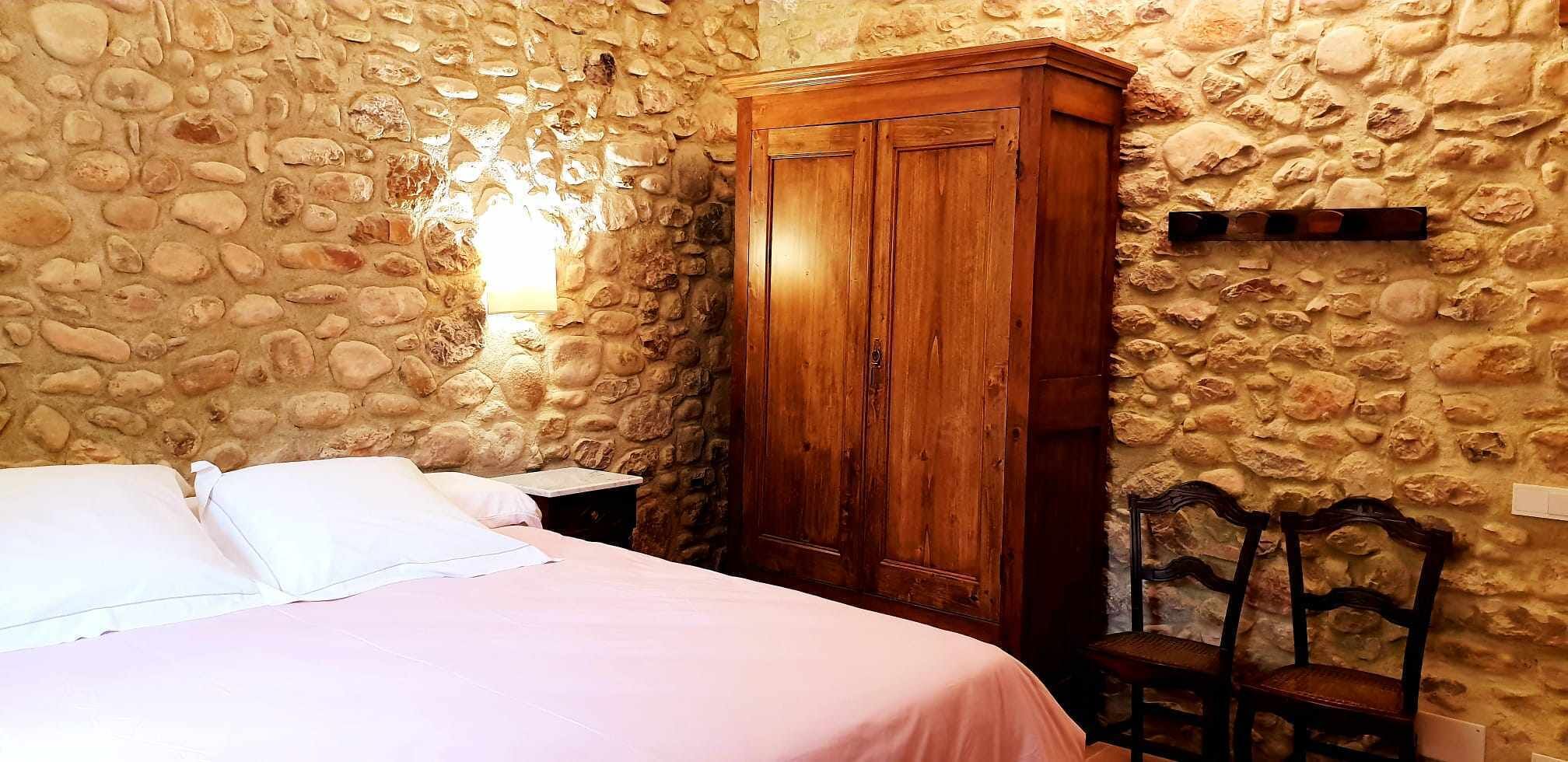 Un dormitorio con pared de piedra, un armario de madera, una cama con ropa blanca y una silla.